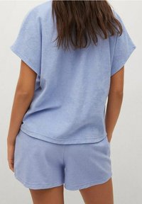 Mujer con cabello castaño largo, vestida con una blusa de manga corta azul claro y pantalones cortos a juego, vista desde atrás contra un fondo liso.