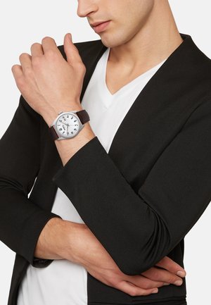 Montre-bracelet argentée avec un bracelet en cuir marron, un cadran blanc, de grands chiffres noirs et un boîtier rond élégant ; portée au poignet d'un homme.