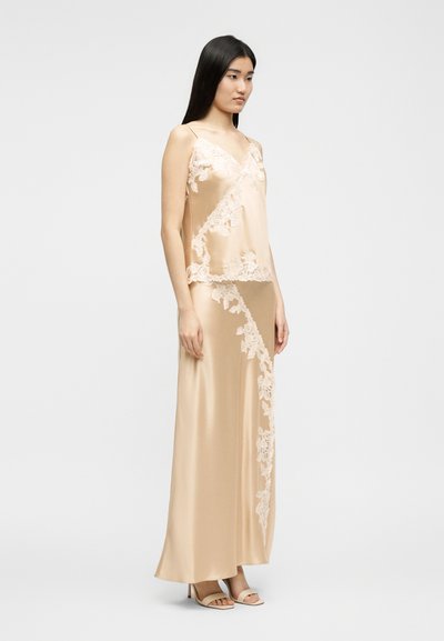 Γυναίκα που φοράει μπεζ σατέν καμπισόλ και μακριά φούστα σετ με λευκές λεπτομέρειες από δαντέλα με λουλούδια, στέκεται μπροστά σε απλό φόντο.