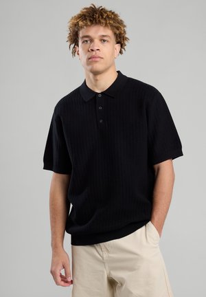 Black short-sleeve polo shirt with a ribbed texture, three-button placket, and a flat collar, paired with light-colored trousers.