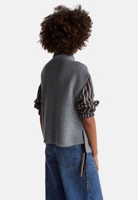 Osoba s kudrnatými vlasy, která má na sobě šedou pletenou vestu bez rukávů přes pruhovanou košili s dlouhými rukávy a modré džíny, stojící zády k bílému pozadí.