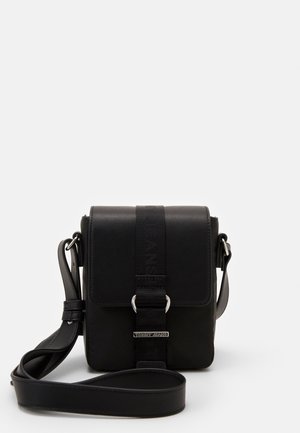 Zwarte faux leren schoudertas met een klep, verstelbare strap, zilveren hardware en een strapdetail met logo-accent. Compact rechthoekige vorm.