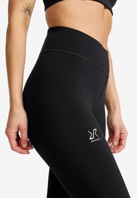 Närbild av en person som bär högmidjade svarta leggings med RVRC-logotyp på låret och en svart sport-bh, som visar en tatuering på vänster arm.