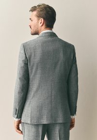 Hallhalline tekstuuriga ülikond, millel on kohandatud lõige, ühe õhutusauguga, nööbitud mansetid ja peen herringbone muster.