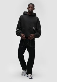 Czarna, oversize'owa bluza z kapturem, z kieszenią z przodu i białymi haftami. W połączeniu z czarnymi spodniami z kontrastowymi szwami i trampkami.