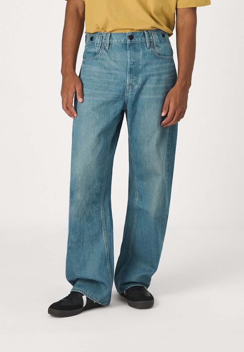 Vaqueros de pierna ancha en mezclilla azul claro, con un diseño clásico de cinco bolsillos, dos botones delanteros y una textura descolorida. Zapatos negros complementan el conjunto.