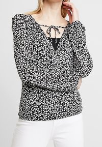 Blusa floreale in bianco e nero con maniche lunghe, scollatura a barchetta con chiusura a legaccio e dettagli arricciati in vita.