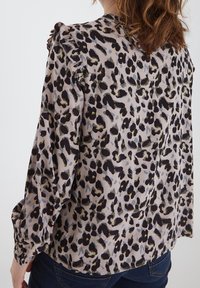 Leopardenmuster-Bluse aus leichtem Material, mit Rüschenakzenten an den Schultern, langen Ärmeln und lockerer Passform. Die dominierenden Farben sind Beige, Schwarz und Gelb.
