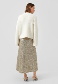 Vrouw in een witte fuzzy trui, gouden pailletten midi-rok, beige hakken, staat met haar rug naar de camera tegen een effen grijze achtergrond.
