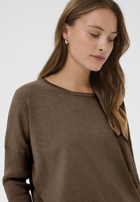 Brauner Strickpullover mit weitem Ausschnitt und gerippten Bündchen, der eine weiche Textur und einen lässigen Schnitt hat. Goldene Creolen werden getragen.