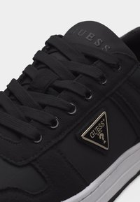 Μαύρο αθλητικό παπούτσι με μαύρα κορδόνια, λευκή σόλα και τριγωνικό μεταλλικό λογότυπο Guess U.S.A. στο πλάι κοντά στα κορδόνια.