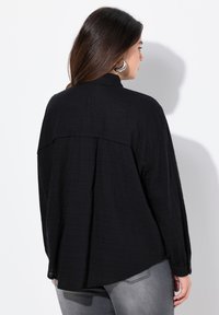 Black, textured long-sleeve shirt with a high neck, relaxed fit, and buttoned cuffs. Notable seam detail across the back.