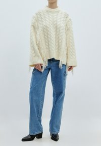 Cremfarbener, oversized gestrickter Pullover mit Zopfmuster, fransigen Kanten, kombiniert mit hellblauen, langen Jeans und schwarzen, hohen Stiefeln.