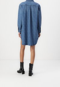 Sininen farkkupaitamekko, jossa toistuva tekstikuvio, pitkät hihat, napilliset hihansuut ja sivuhalkiot. Yhdistetty mustien nilkkureiden kanssa.