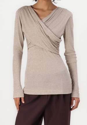 Mujer con blusa beige de manga larga, estilo cruzado con escote en V y detalle fruncido en el lateral, combinada con pantalones sueltos marrón oscuro.