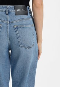 Gaiši zili džinsa džinsi ar vaļēju fitu, ar divām aizmugurējām kabatām un melnu ādas etiķeti "HUGO" pie jostasvietas.