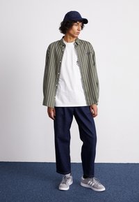 Groen en wit gestreepte button-up shirt over een plain wit T-shirt, gecombineerd met donkere spijkerbroeken en grijze sneakers, en een pet.