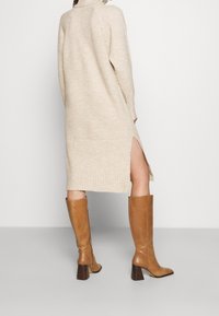 Beige ribbed knit dress with a high neck and side slits, paired with brown knee-high heeled boots featuring a square toe and textured accents.
