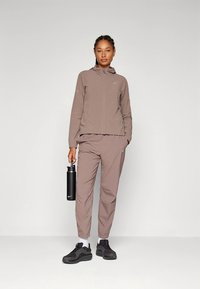 Svetlohnedá športová bunda a nohavice s hladkou textúrou, so zipsom vpredu a logom Nike. Čierna fľaša na vodu a tmavé tenisky.