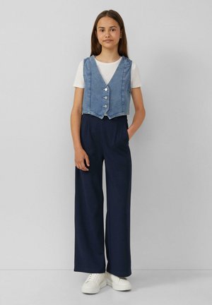 Gekürzter Jeansmantel mit drei Knöpfen über einem weißen T-Shirt, kombiniert mit dunkelblauen, weit geschnittenen Hosen und weißen Sneakers.