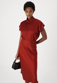 Abito midi in satin rosso con collo alto, maniche a volantino corte, vita aderente e texture liscia. Accompagnato da una piccola borsa nera.