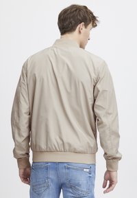 Lysebeige bomberjakke med en glatt, glansfull tekstur, ribbede mansjetter og nederkant, og en avslappet passform, båret over blå jeans.