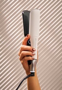 Main tenant un lisseur de cheveux ghd chroma max noir et blanc avec des ongles manucurés couleur cuivre sur un fond texturé.