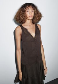 Persona con capelli ricci che indossa un top senza maniche color marrone scuro a scollo a V e una gonna coordinata, in piedi di fronte a uno sfondo chiaro e semplice.