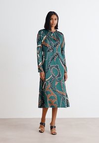 Grünes Midi-Kleid mit langen Ärmeln, das ein Kettenmuster in Brauntönen und Creme zeigt; aus glattem Stoff gefertigt, mit einer lässigen Passform.