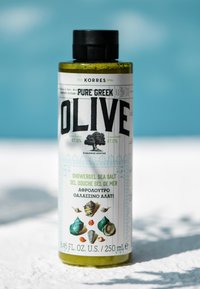 Bouteille verte de gel douche avec un capuchon noir, étiquetée "Pure Greek Olive." Présente des graphiques de coquillages et du texte en noir et vert.