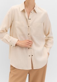 Chemise beige à boutons avec deux poches poitrine, manches longues et coupe légèrement ample. Dotée de boutons en métal et d'une texture lisse.