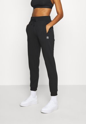 Persoon draagt zwarte Adidas joggingbroek, zwart cropped top, witte sokken en witte sneakers, staat met handen in de zakken.