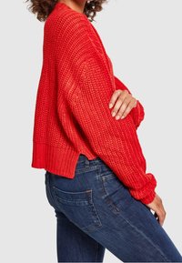Femme portant un pull en maille épaisse rouge vif à manches longues et un jean en denim bleu, debout de profil avec un bras croisé.