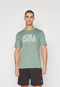 Grünes Baumwoll-T-Shirt mit dem weißen Grafikaufdruck "PUMA Sports Club". Kurze Ärmel, Rundhals, weiche Textur. Model trägt außerdem schwarze Shorts.