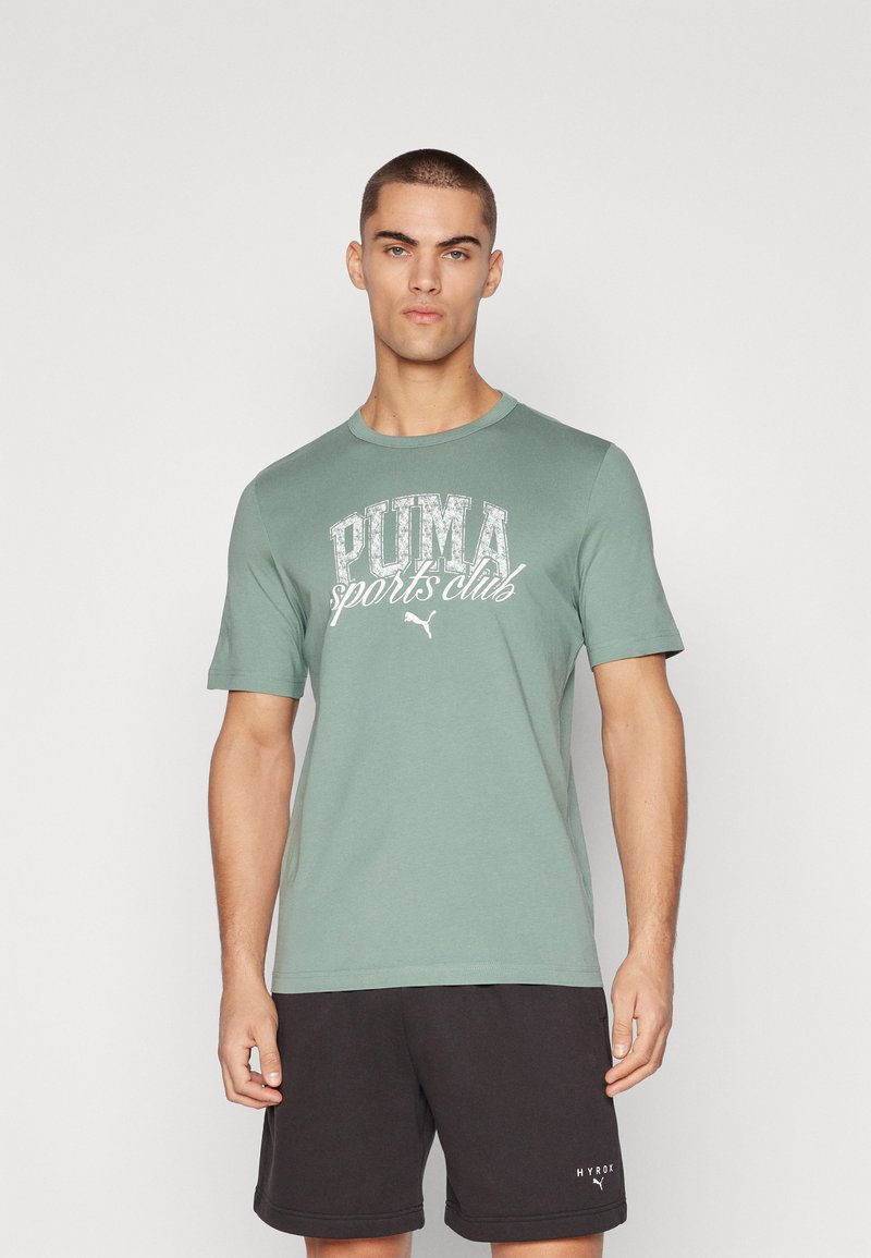 Grünes Baumwoll-T-Shirt mit dem weißen Grafikaufdruck "PUMA Sports Club". Kurze Ärmel, Rundhals, weiche Textur. Model trägt außerdem schwarze Shorts.
