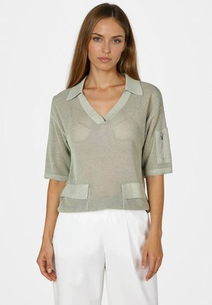 Woman wearing a light green knitted short-sleeve top with collar and zipper pocket on sleeve, paired with white pants, standing still.