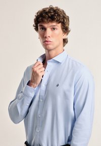 Camisa de manga larga azul claro de algodón, con cuello abotonado y un pequeño logo oscuro en el lado izquierdo del pecho. Textura suave.