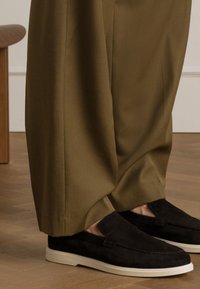 Black suede slip-on shoes with a white rubber sole, paired with olive green, loose-fitting trousers. Smooth textures and clean lines are visible.