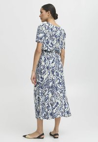 Robe midi à motif floral en tons blancs et bleus, avec des manches courtes, une taille cintrée et une jupe fluide. Portée avec des ballerines noires et beiges.