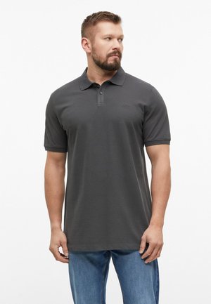Mann mit kurzen braunen Haaren und Bart, der ein dunkelgraues kurzärmliges Poloshirt und blaue Jeans trägt, schaut nach links.