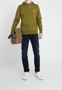 Olivgrön hoodie med dragsnören, känguruficka, logotyp på bröstet; i kombination med mörk denimjeans och vita sneakers, hållande en brun väska.