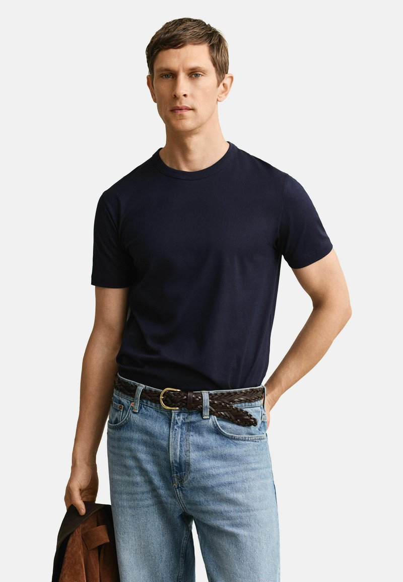 Hombre vistiendo una camiseta azul marino y jeans azul claro con un cinturón trenzado marrón, sosteniendo una chaqueta marrón en su mano izquierda.