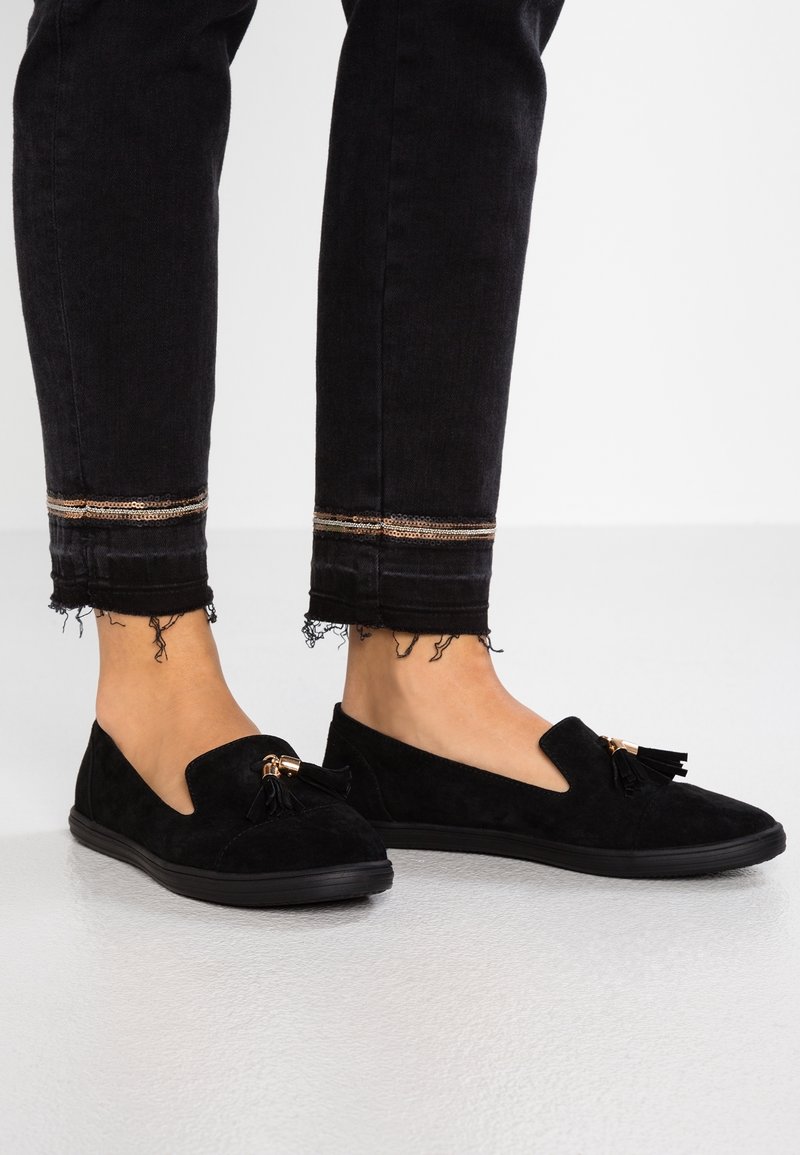 Jambes portant un jean slim noir aux ourlets effilochés et ornés d'une bordure à sequins dorés, assorti à des mocassins en daim noir avec des décorations à pampilles.