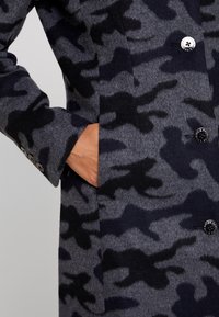 Grijze wollen jas met zwart camouflagepatroon, voorzien van een enkele rij knopen en zijzakken. Gestructureerde stof en een op maat gemaakte pasvorm.