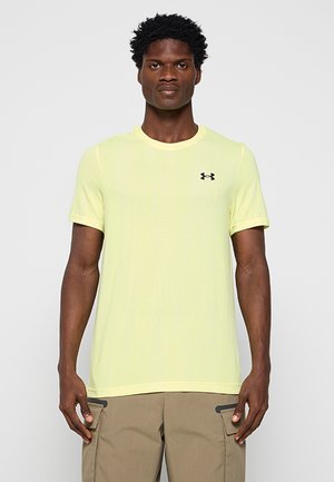 Mężczyzna ubrany w cytrynowo-żółtą koszulkę Under Armour z krótkim rękawem oraz beżowe spodnie typu cargo, stojący na jednolitym jasnoszarym tle.