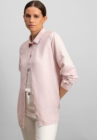 Camicia button-up rosa chiaro in un tessuto leggero, con colletto, maniche lunghe e polsini arrotolati. Abbinata a pantaloni bianchi.