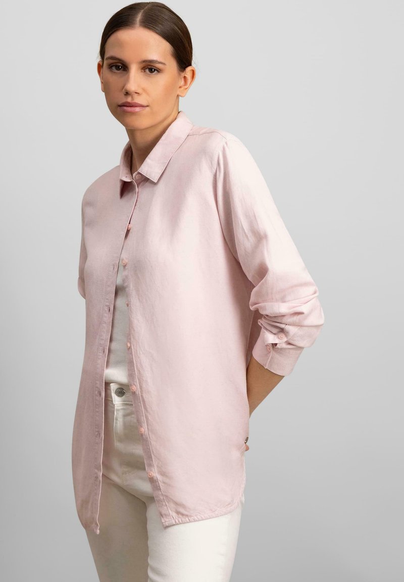 Camicia button-up rosa chiaro in un tessuto leggero, con colletto, maniche lunghe e polsini arrotolati. Abbinata a pantaloni bianchi.