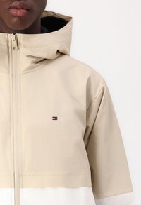 Beżowa kurtka z kapturem o teksturowanej powierzchni, z białym dolnym panelem i małym logo w kolorach czerwonym, białym i niebieskim na piersi.