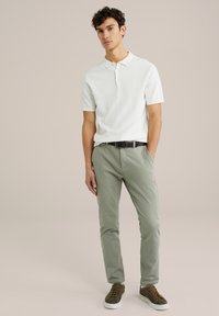 Cămașă polo albă cu dungi, pantaloni verzi deschiși, curea neagră, adidași maro. Model cu părul creț stând pe un fundal neutru.