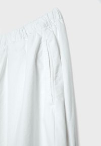 Pantalon à taille élastique blanche avec une poche fendue sur le côté droit, confectionné dans un tissu doux et léger avec des détails de surpiqûres visibles.